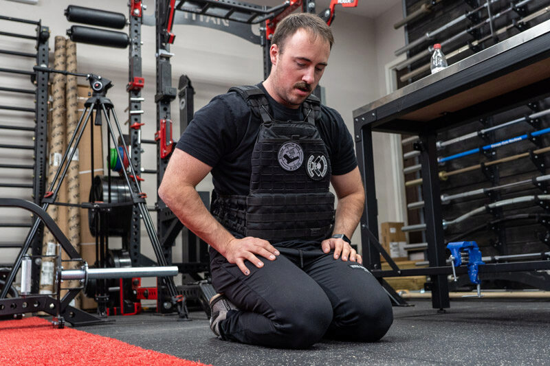 A man sitting on his knees with his hands on his thighs, wearing the black 5.11 TacTec Plate Carrier weighted vest.