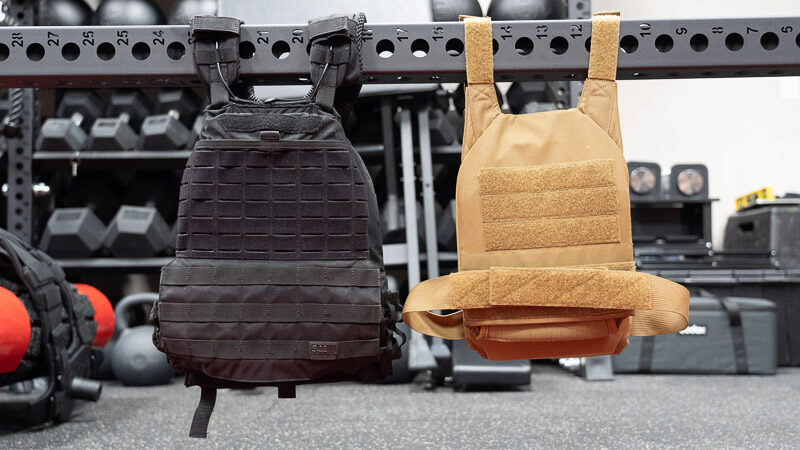 The dark tan Rogue Plate Carrier weighted vest hanging on a metal bar to the right of a black weighted vest