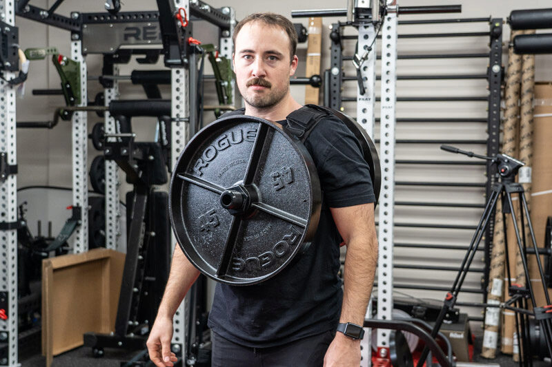 A man standing in front of REP Fitness strength training equipment, while wearing the Kensui EZ-VEST Plate-Loadable Weight Vest with two 45 lb. Rogue Fitness weight plates attached.