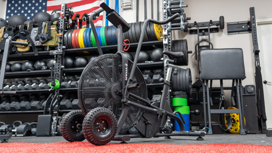The Titan Fitness Air Bike is displayed inside of Coop's garage gym. The bike is all black and the carpet is red.