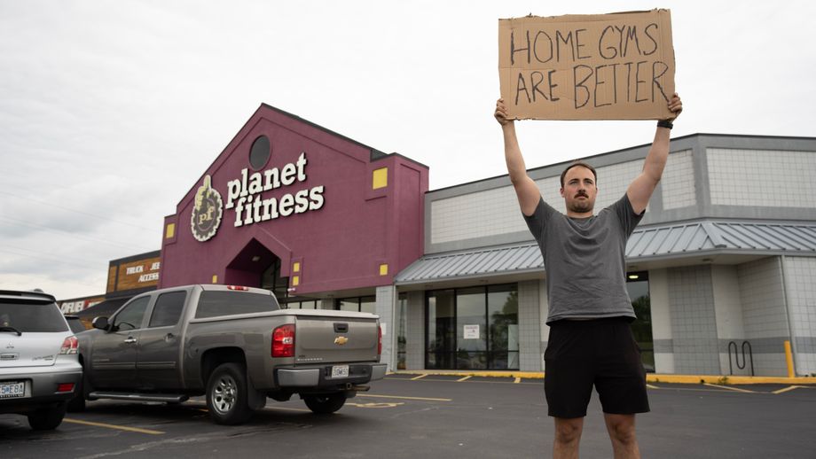 Coop holding a sign that says "Home gyms are better" in front of a Planet Fitness