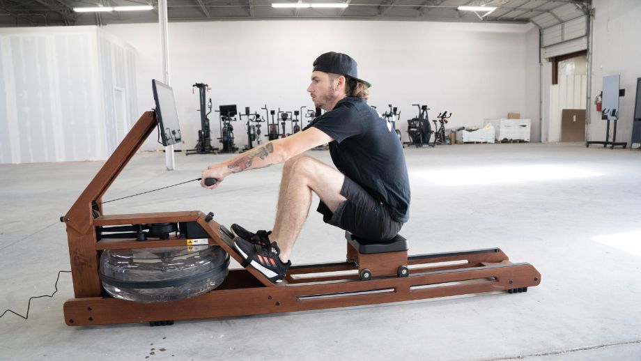 Man in shorts, a t-shirt, and a baseball hat using the Ergatta Rower in a warehouse