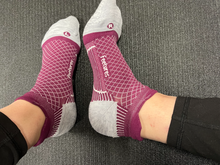 A close-up of feet wearing vibrant purple Feetures Plantar Fasciitis Relief Socks with grey toes, heels, and a dotted grid pattern, highlighting their design for enhanced comfort and support.