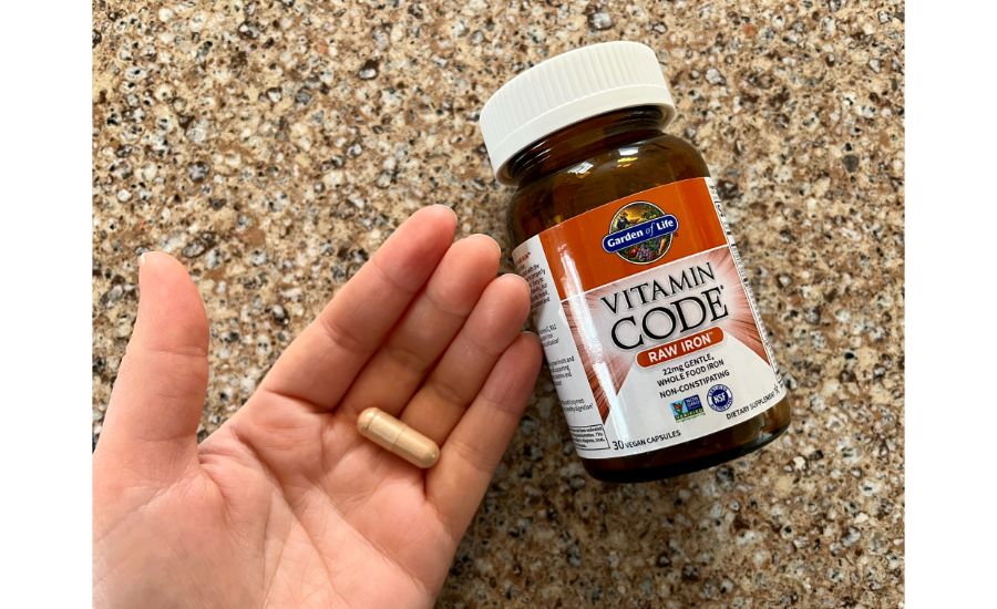 Close-up of a hand holding a single large capsule next to a brown bottle with red labeling of the Garden of Life Vitamin Code raw iron supplements.