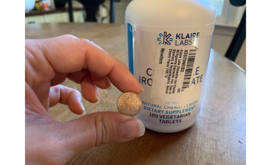 Close-up of a hand holding a large and round brown chewable tablet next to a white bottle of Klaire Labs Chewable Iron Chelate supplements.