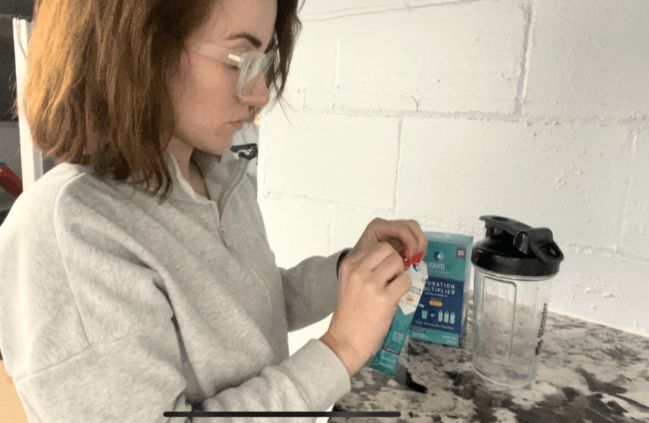 A person wearing glasses and a gray sweatshirt is preparing a drink at a counter. They are opening a packet beside a clear shaker bottle. A blue box of Liquid I.V. hydration packets is on the counter nearby.