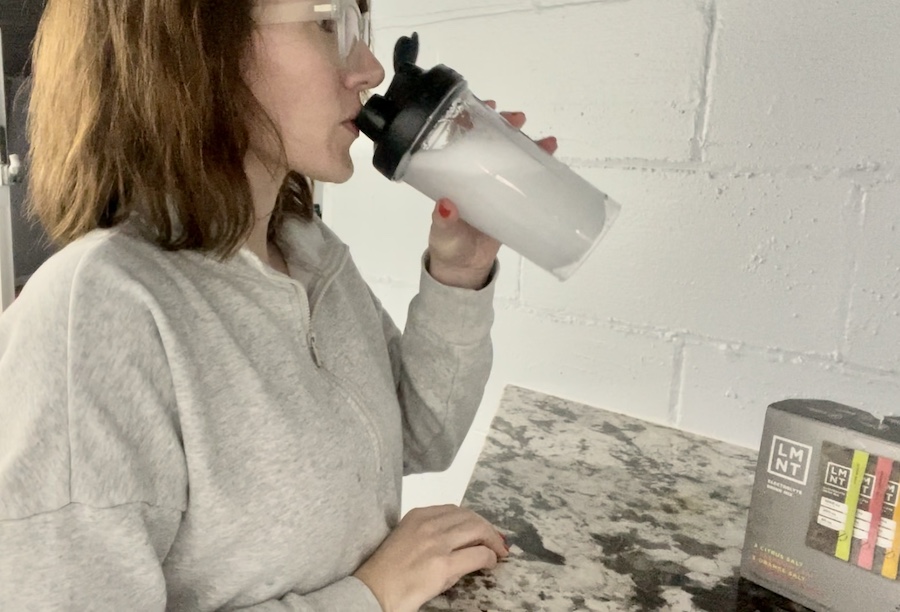 A person in a gray sweatshirt drinks from LMNT a shaker bottle while standing at a countertop. There is a box of LMNT electrolyte packets on the counter. The wall in the background is made of light-colored bricks.