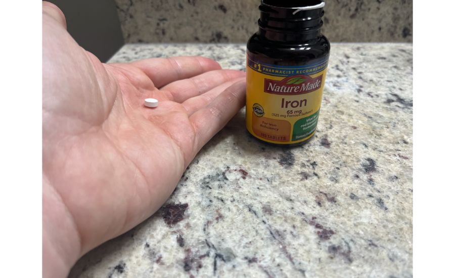 A hand holding a single tablet next to a brown bottle with colorful labeling of Nature Made Iron 65mg supplements.