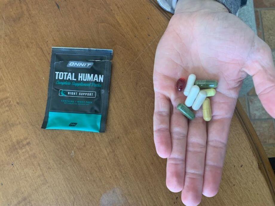 Close-up of a hand holding a handful of colorful supplement capsules and softgels, next to a torn open packet of Onnit Total Human supplements.