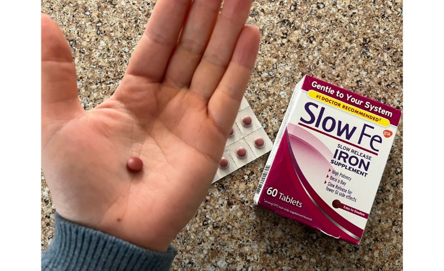 Close-up of a hand holding a single small red tablet next to an aluminum sealed pack of more tablets and a box of Slow Fe Slow Release 45mg Iron supplements.