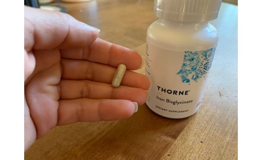 Close-up of a hand holding a single capsule of the Thorne Iron Bisglycinate dietary supplement, with the white and blue bottle sitting in the background.