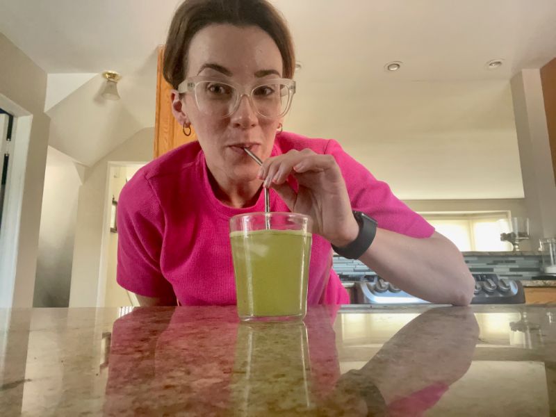 A woman drinking 8greens Greens Powder out of a glass with a metal straw.
