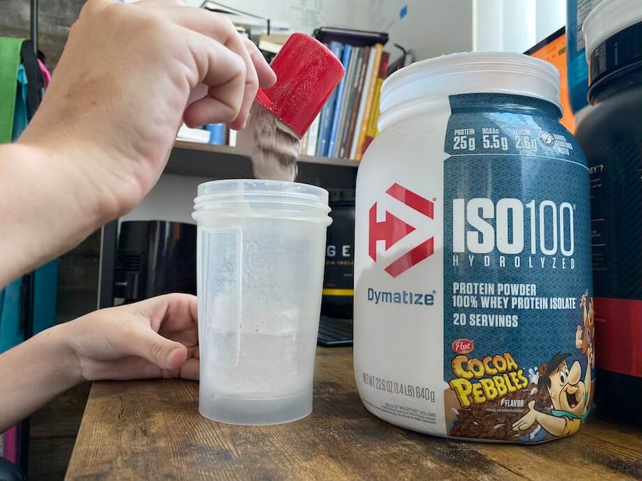 A person pouring a scoop of Dymatize ISO 100 into a shaker bottle