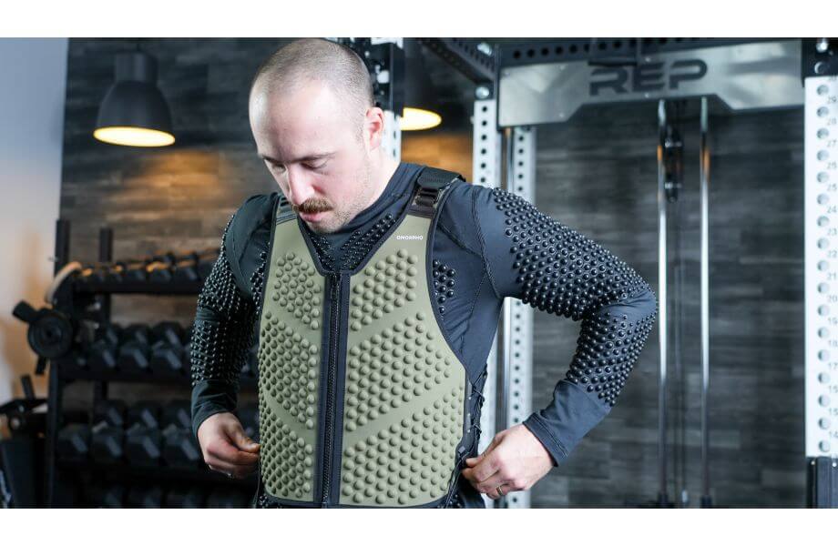 A man showcases his workout in a gym, wearing a green OMORPHO G-Vest+ and weighted black long-sleeved shirt for added resistance.