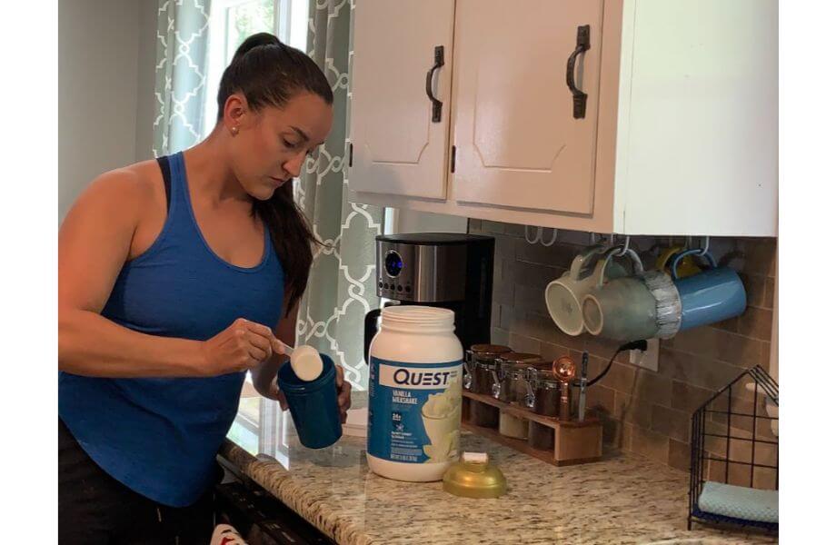 A woman in a blue tank top scooping Quest Protein out of the container into a blue shaker cup