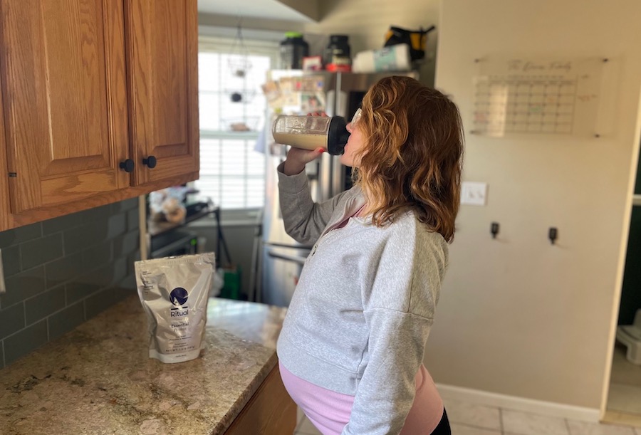 A pregnant woman standing in a kitchen drinking Ritual Essential protein powder for pregnancy out of a clear shaker bottle