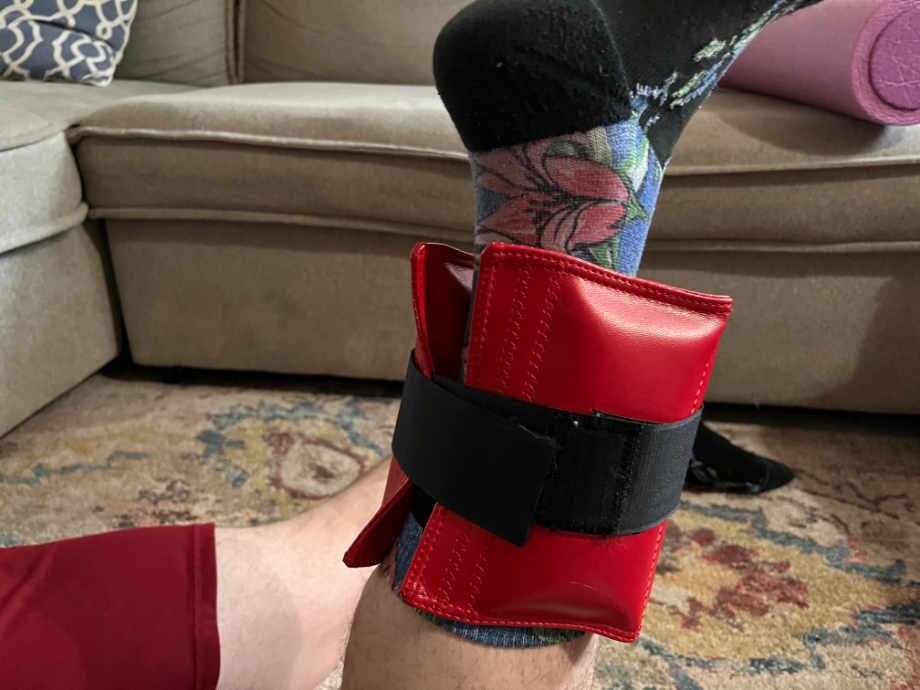 A Close-up shot of a man's left ankle while wearing The Cuff ankle weight in red, strapped on in prone position