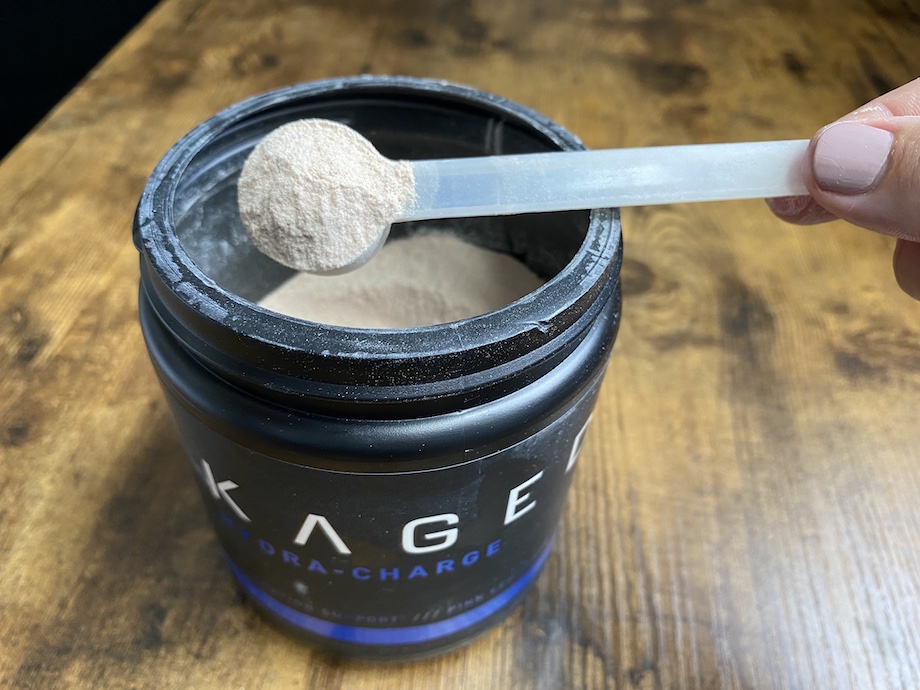 A hand holding a clear plastic scoop filled with beige powder above an open black container labeled with KAGE on a wooden surface.