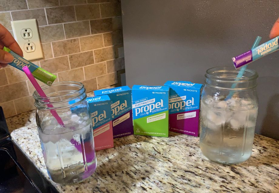 Two jars of water on a kitchen counter, each being stirred with straws. In the background, there are boxes of Propel powder drink mix in different flavors, and packets of the mix are being poured into the jars.