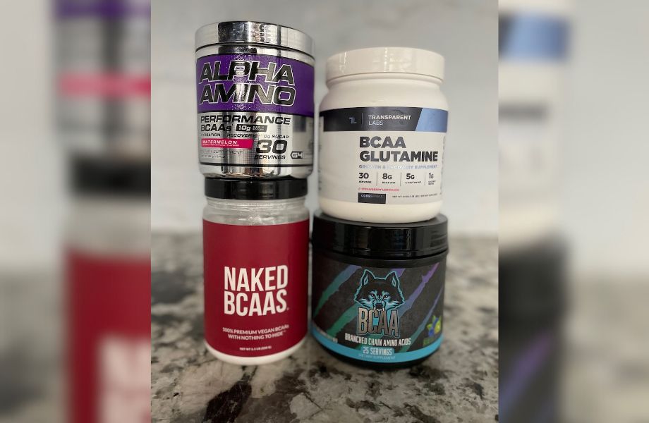 Four containers of dietary supplements are stacked. Labels read: Alpha Amino, BCAA Glutamine, Naked BCAAs, and another BCAA product with a wolf design. They are placed on a marble surface.
