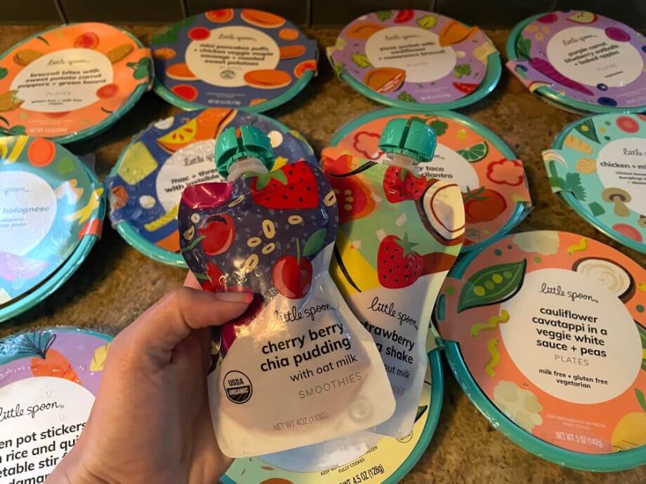 A person holds two smoothie pouches in front of an array of plates from Little Spoon