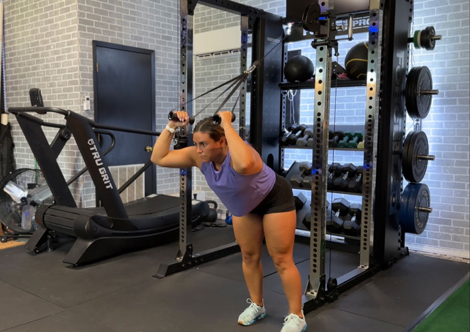 A person in a purple shirt and black shorts performs a cable tricep pushdown exercise in a gym. Nearby are weights, a treadmill, medicine balls, and wall pads. The walls are gray with a pattern.