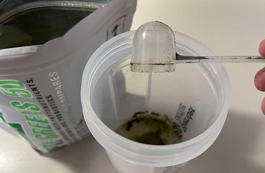 Dumping a scoop of Opti Greens 50 into a clear shaker cup