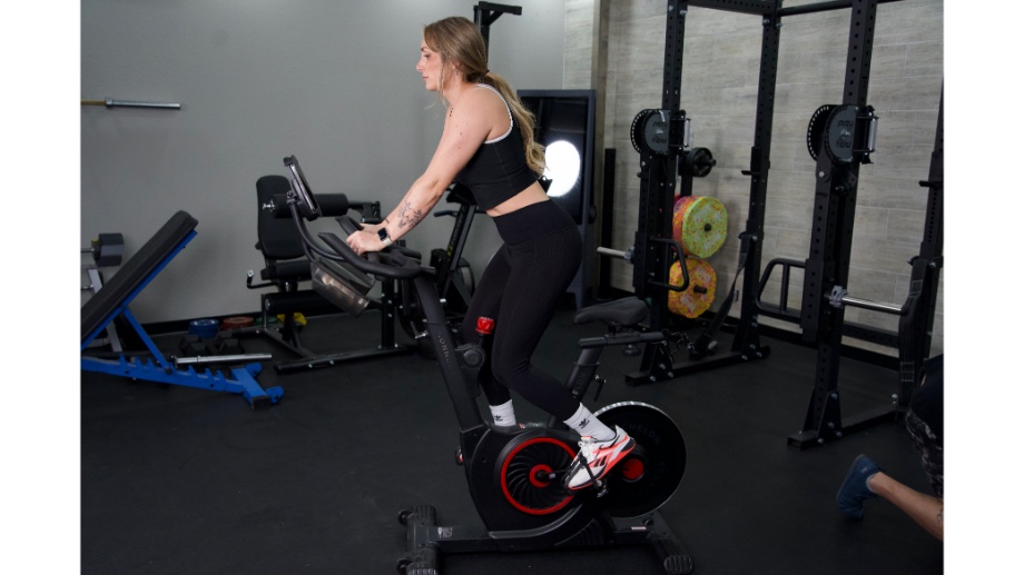 Woman standing up riding a Peloton alternative, the echelon ex5 bike with other gym equipment in the background