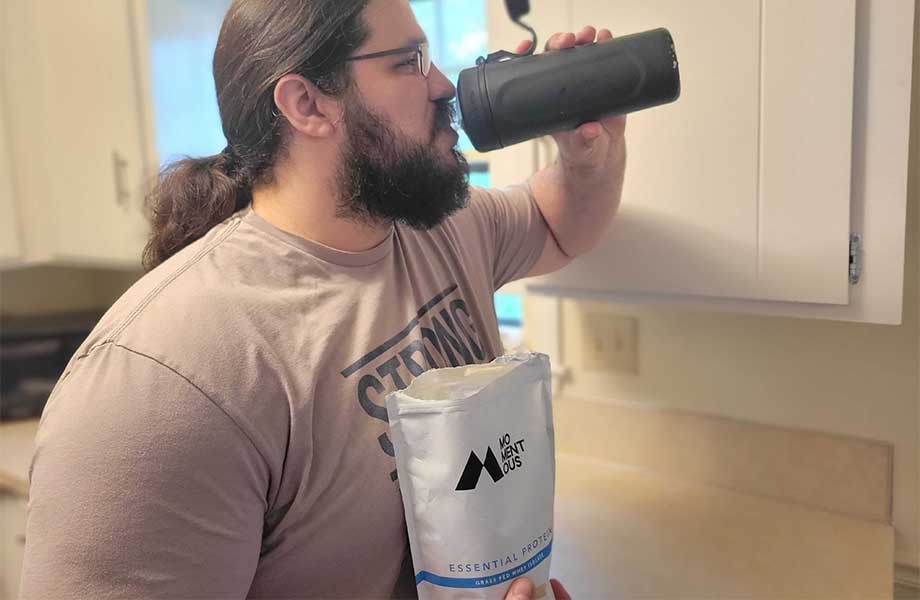 A man with a ponytail and beard drinking out of a black shaker bottle while holding a bag of Momentous Whey protein powder