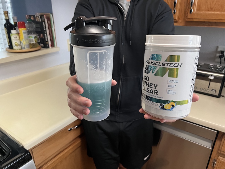 A person holds out a shaker bottle containing a blue liquid and a container of MuscleTech Clear Whey Isolate