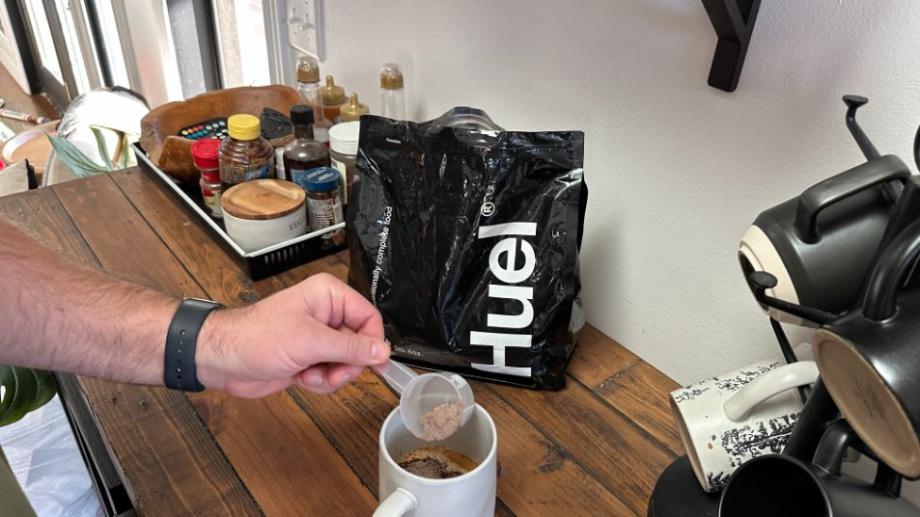 Someone adding a scoop of Huel Black Edition to their mug of coffee. The black Huel bag is in the background.