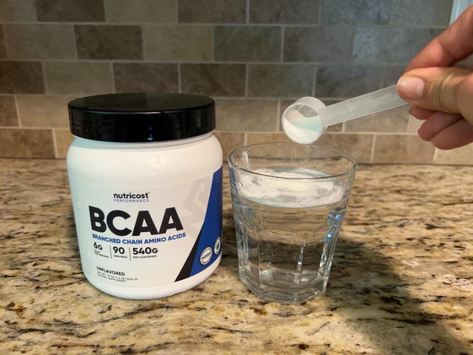 A hand holds a scoop of white powder over a glass of water. Next to the glass is a container of Nutricost BCAA powder. The background features a kitchen countertop and a tiled backsplash.