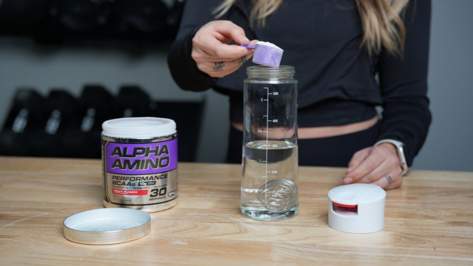 A person in a black long-sleeve top is scooping powder from a container labeled Alpha Amino Performance BCAAs into a clear water bottle with measurement markings. The bottle sits on a wooden table beside the container lid.