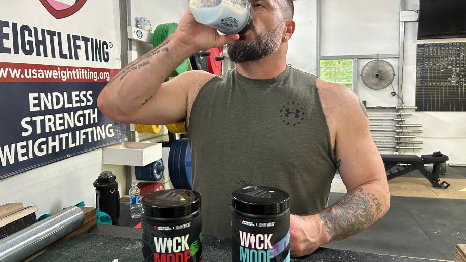 A man in a gym drinks from a shaker bottle. He wears a sleeveless shirt and is surrounded by weightlifting equipment. Two supplement containers labeled WCKD MODE are on a nearby table. A wall sign reads Endless Strength Weightlifting.