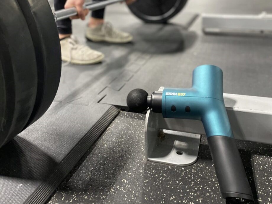 A person deadlifts with the Ekrin Athletics B37 massage gun placed in front of them, resting on the feet of a squat stand