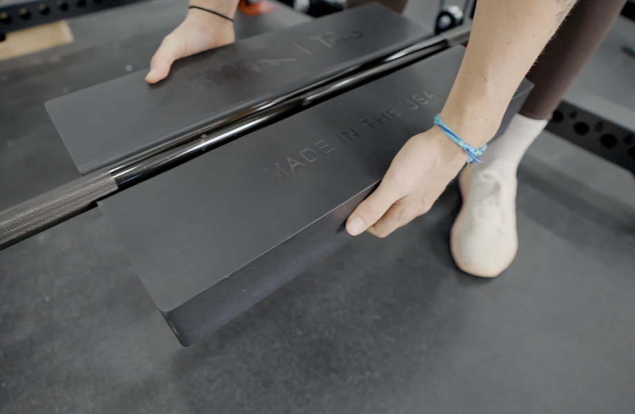 A person holds an AbMat Pivot Pad to show how a groove on the back is used to clip it onto a barbell.