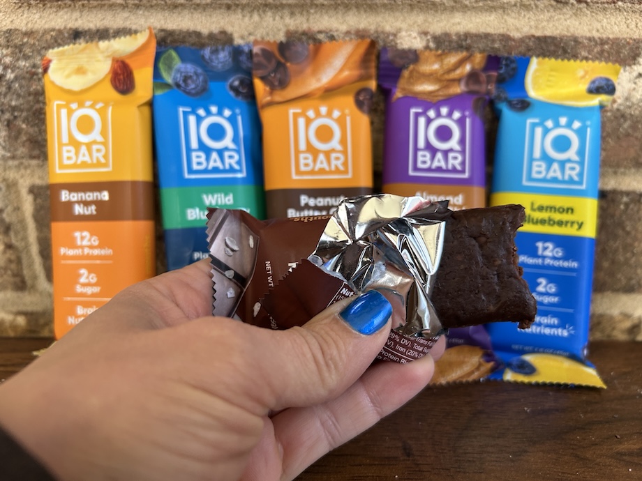 Woman holding an open IQBAR Protein Bar next to several other flavors.