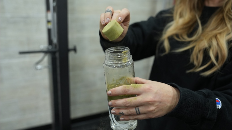 A woman pours a scoop of AG1 greens powder into a shaker bottle