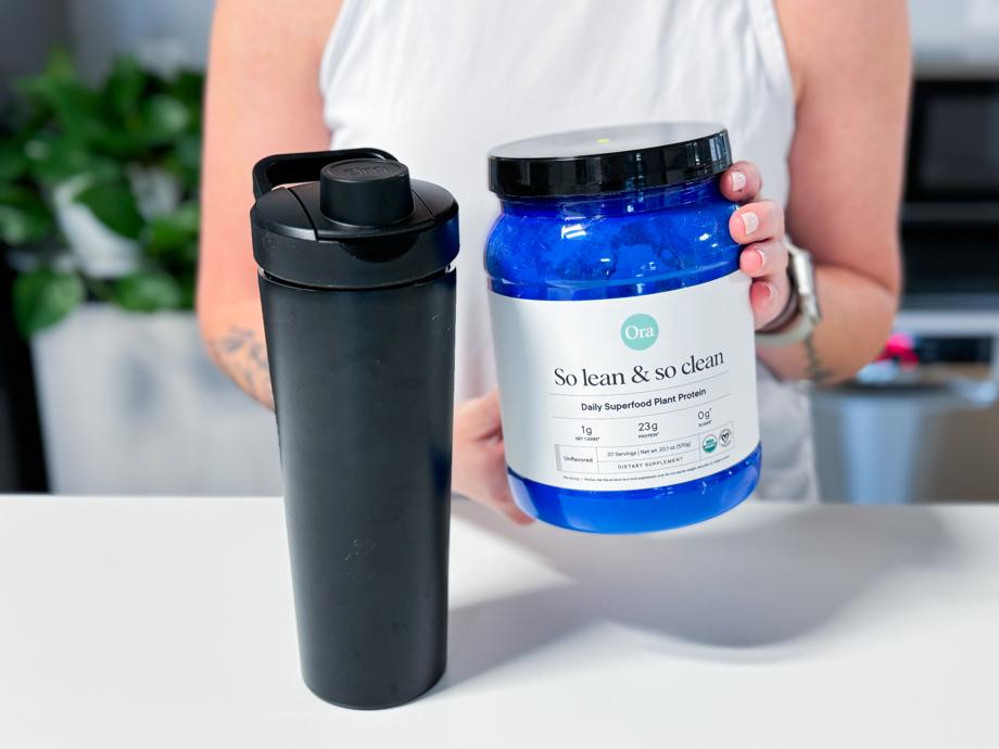 Holding a tub of Ora Organics So Lean & So Clean Plant Based Superfood Protein