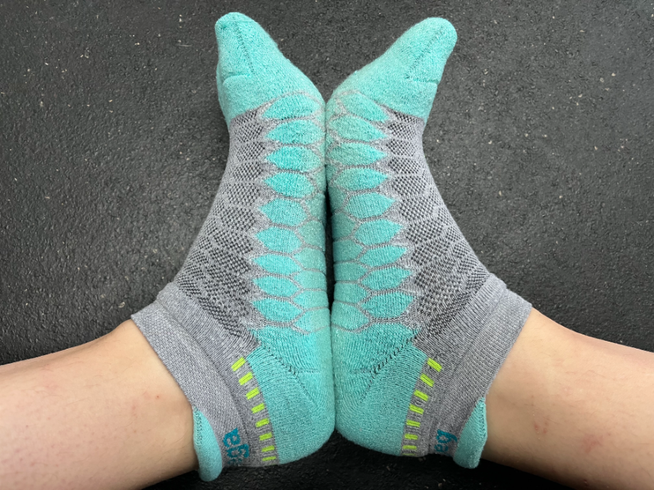 Close-up of a pair of feet in blue and grey Balega Silver No-Show Compression-Fit Running Socks with green heel accents, emphasizing athletic design and fit.
