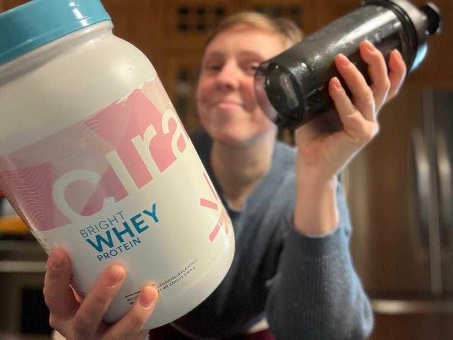 A woman is smiling while holding out a shake and container of Cira Nutrition Bright Whey Protein.