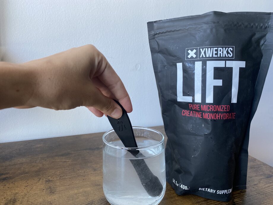 A hand is shown stirring XWERKS Lift creatine into a glass of water