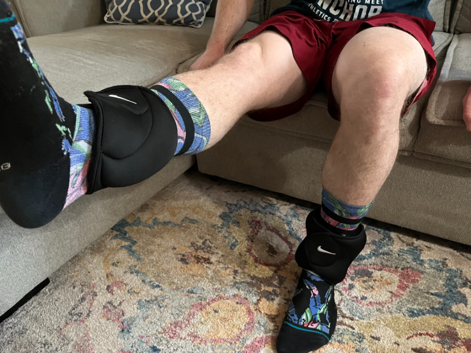 A man performing a seated quad extension while wearing the black Nike ankle weights and sitting on a sectional couch