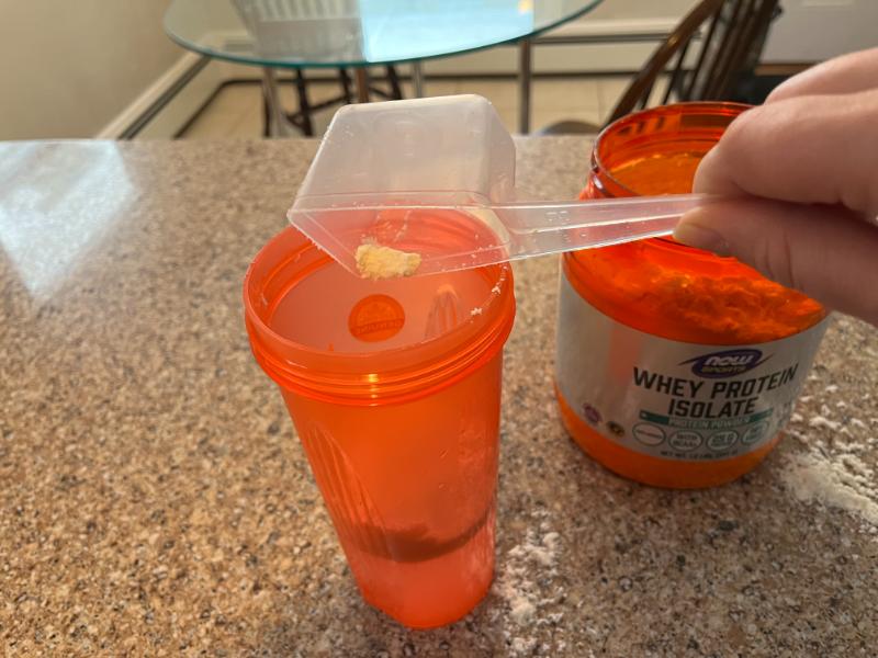 Closeup of a hand mixing a batch of NOW Sports Whey Protein Isolate in an orange shaker cup