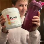 A smiling woman holds up a shaker cup and a container of RAW Nutrition Pump pre-workout.