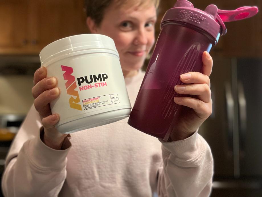A smiling woman holds up a shaker cup and a container of RAW Nutrition Pump pre-workout.