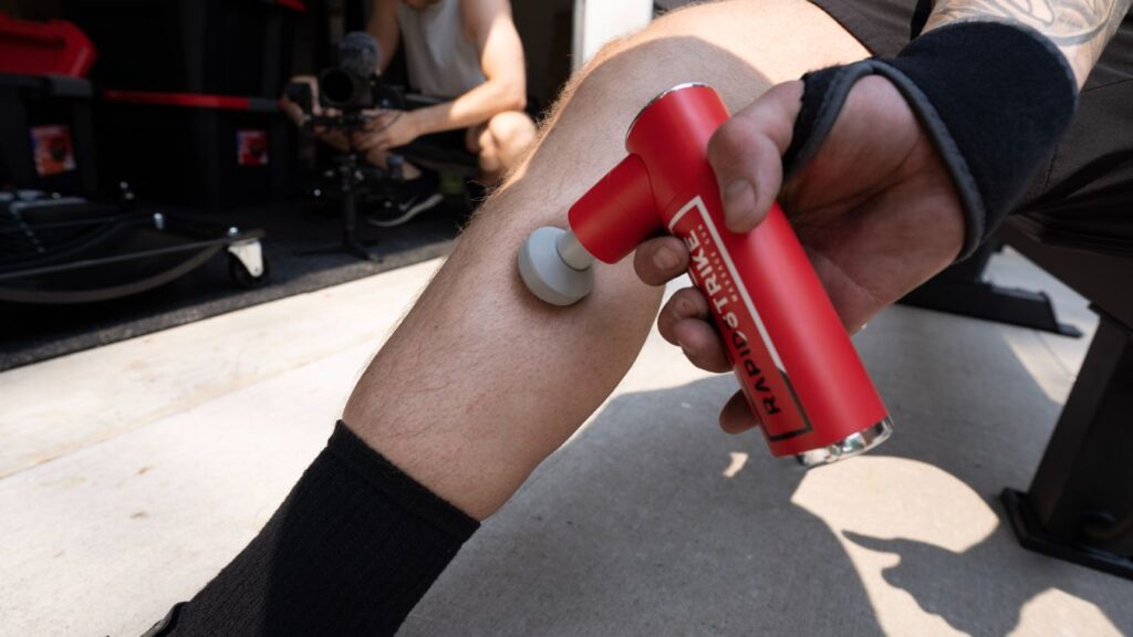 Sitting outside of a garage gym using the REP Rapidstrike massage gun on calf muscle with the round attachment.