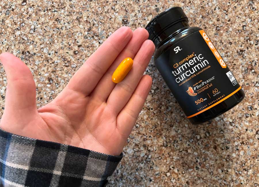 A hand holding a Sports Research turmeric curcumin pill next to the supplement bottle