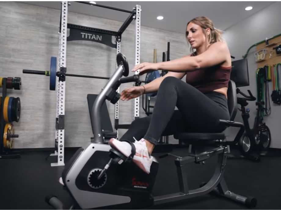 A person is shown reaching for the handles on the Sunny Health and Fitness Essential Recumbent Bike.