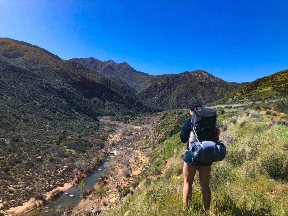 A woman backpacking along a river with mountains in the background